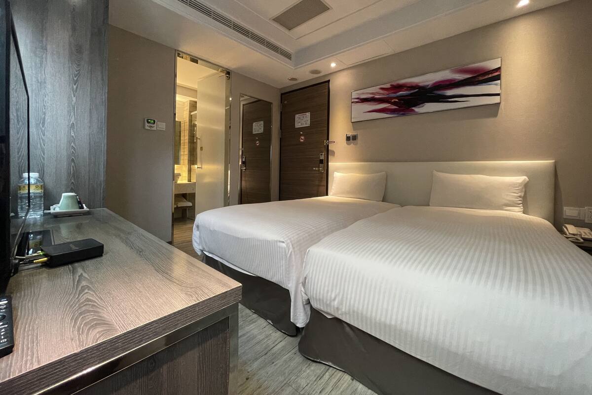 Une chambre d’hôtel avec deux lits, un bureau avec un ordinateur et une salle de bain visible à travers une porte ouverte.