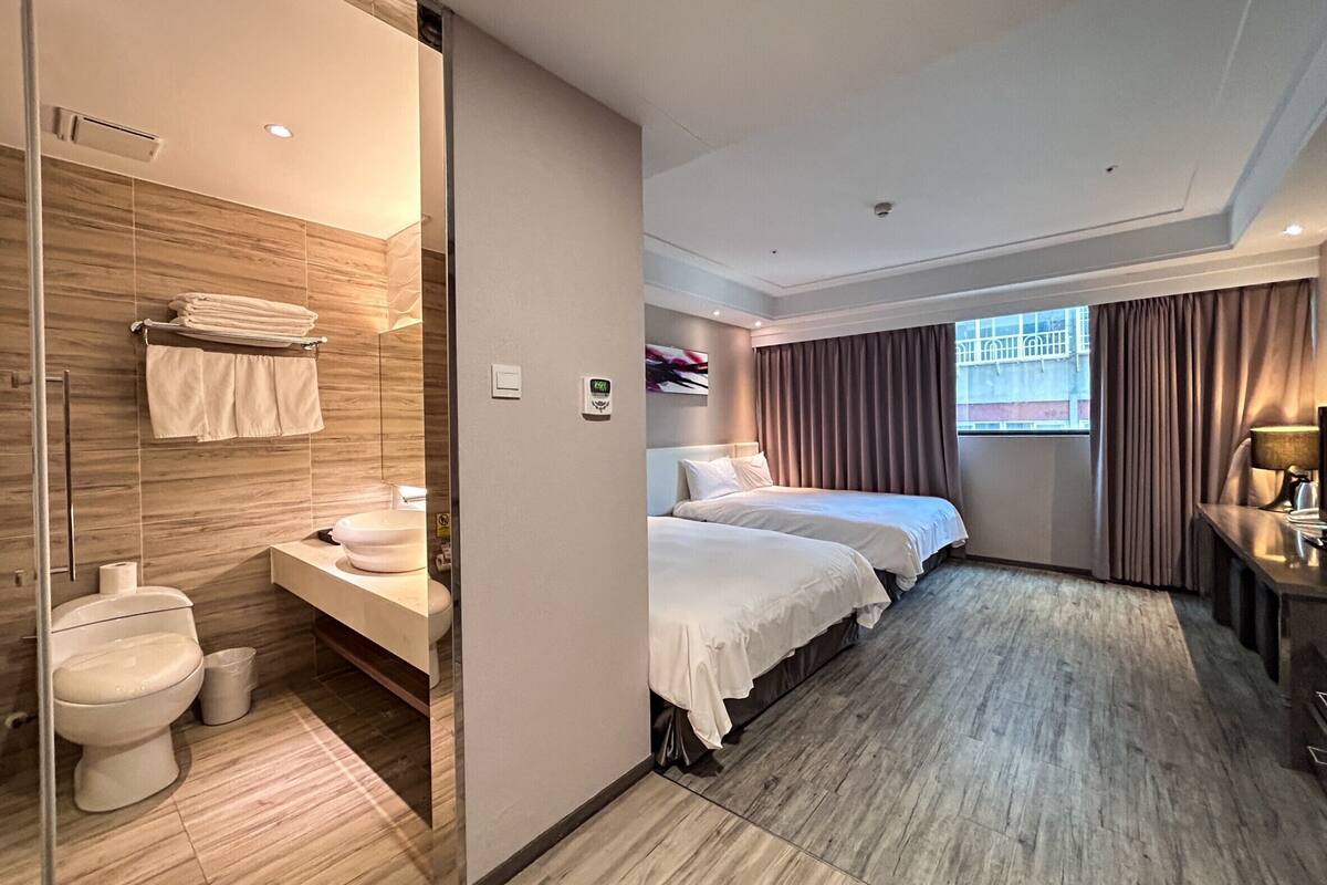 Une chambre d’hôtel avec un plancher en bois, deux lits, une salle de bain avec un lavabo et des toilettes, et une fenêtre avec des rideaux.