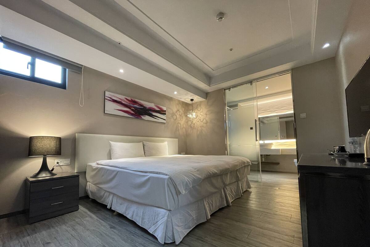 Une chambre moderne avec un grand lit, des tables de chevet, un bureau et une salle de bain visible à travers une porte en verre.