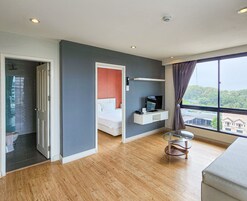 Una habitación de hotel moderna con piso de madera, una cama con ropa blanca, un televisor sobre un estante blanco y un amplio ventanal con vistas a edificios y árboles.