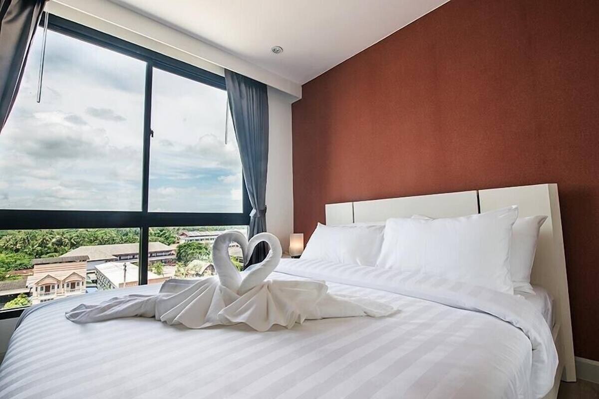 A hotel room with a large bed, a heart-shaped towel decoration, a nightstand with a lamp, and a view of buildings and greenery through the window.