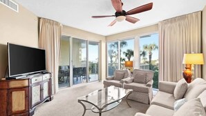 A spacious living room with a flat-screen TV, a glass coffee table, and a ceiling fan.