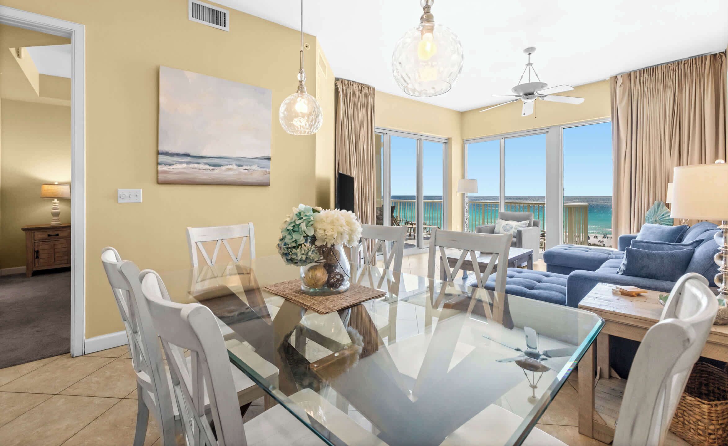 A spacious living area with a glass table, white chairs, a blue sofa, and a view of the ocean.