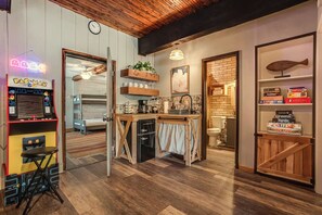 A compact kitchen with wooden flooring, a small dining area with a bench, a neon sign, and a vintage arcade machine.