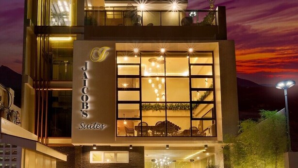 Un hôtel moderne doté d’une façade en verre, d’un espace extérieur aménagé et d’une enseigne indiquant « JAICOB’S Suites ».