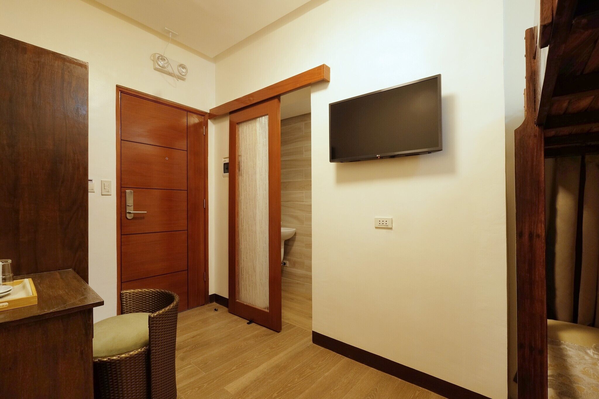Una habitación con un armario de madera, un televisor montado en la pared, una silla de mimbre y una mesita con un vaso y un plato.