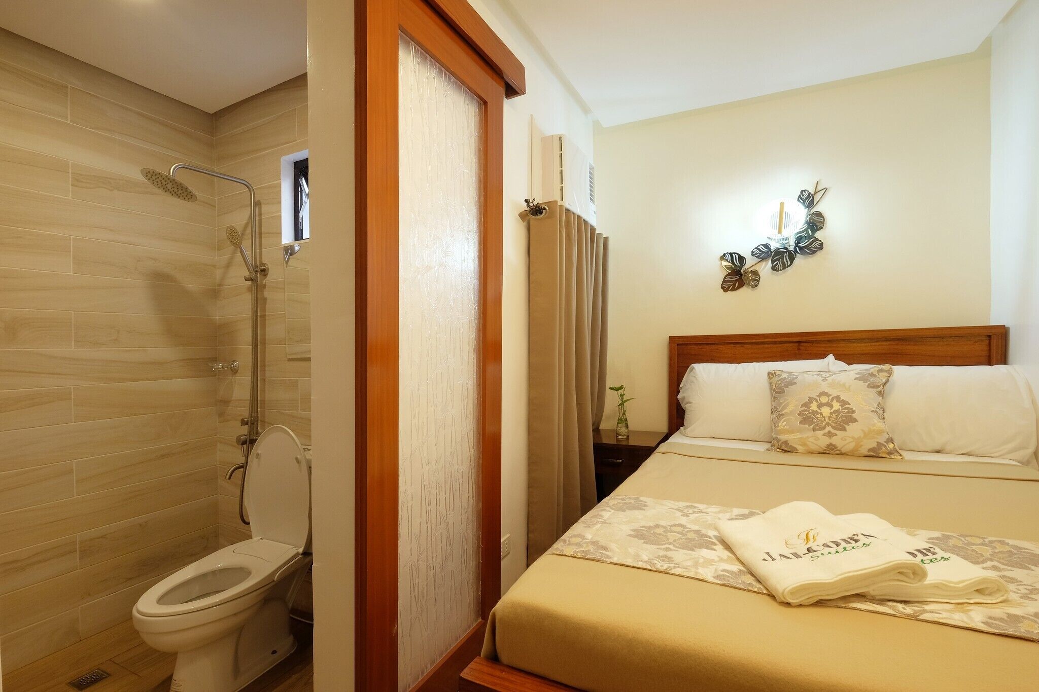 A hotel room with a wooden floor, a white toilet, a shower, a bed with a beige bedspread and a pillow, and a wall clock.