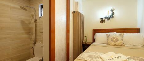 A hotel room with a wooden floor, a white toilet, a shower, a bed with a beige bedspread and a pillow, and a wall clock.