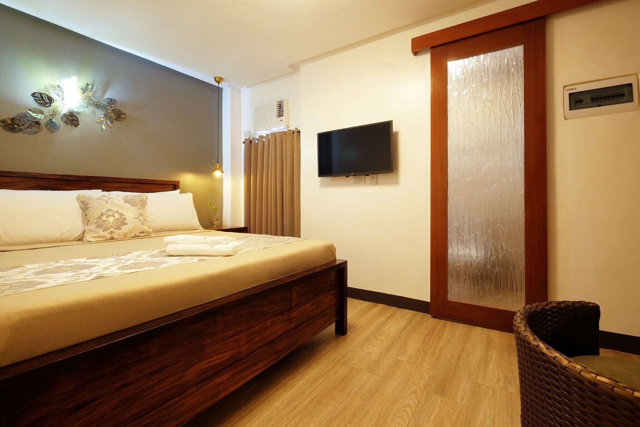 A hotel room with a bed, a television, a wall-mounted air conditioner, and a wicker chair.