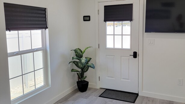 Un intérieur moderne avec une porte blanche, deux fenêtres équipées de stores, une plante en pot et un banc en cuir noir.