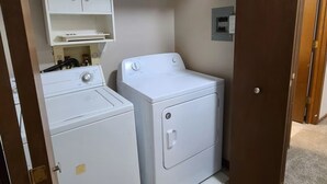 Une buanderie équipée d’un lave-linge et d’un sèche-linge, avec une étagère au-dessus du lave-linge et une porte donnant sur une autre pièce.
