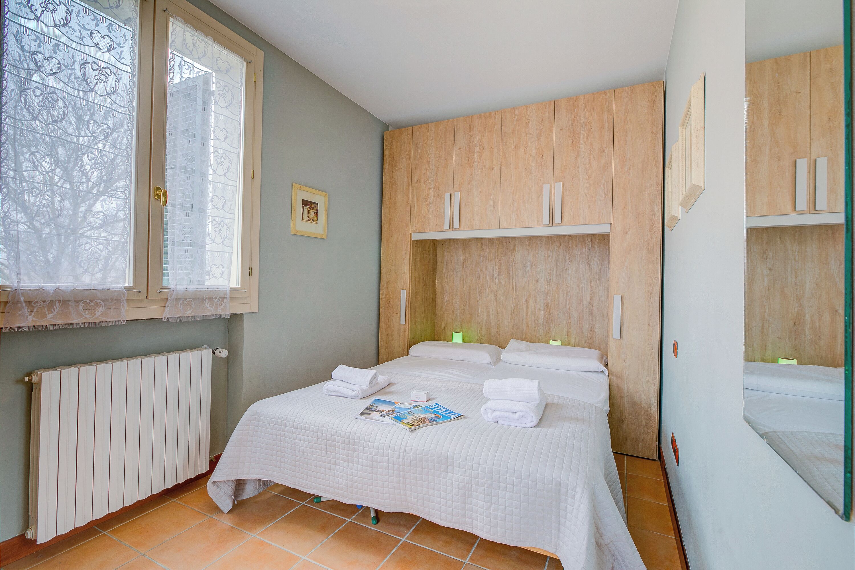 A hotel room with a bed, a window with a view of trees, a wooden wardrobe, and a radiator.