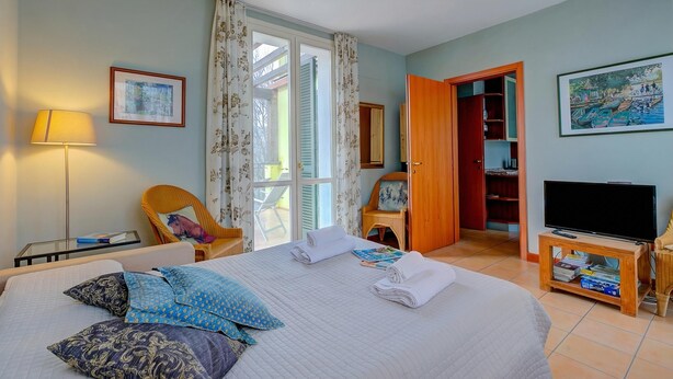 A hotel room with a bed, a chair, a TV, a bookshelf, and a door leading to a balcony. - Porto Letizia Orchidea 3, Porlezza, Italy (Porlezza)