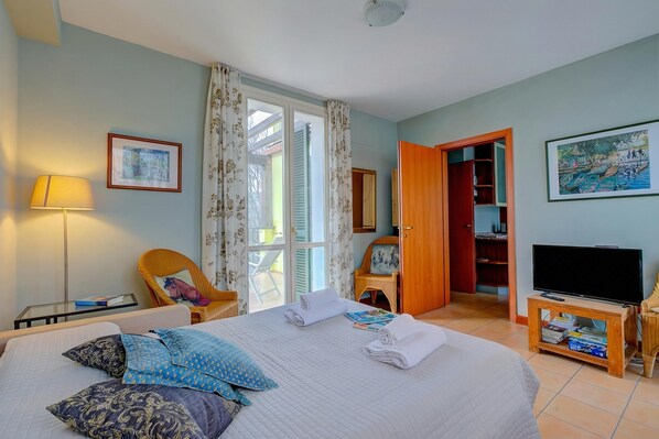 A hotel room with a bed, a chair, a TV, a bookshelf, and a door leading to a balcony. - Porto Letizia Orchidea 3, Porlezza, Italy (Porlezza)