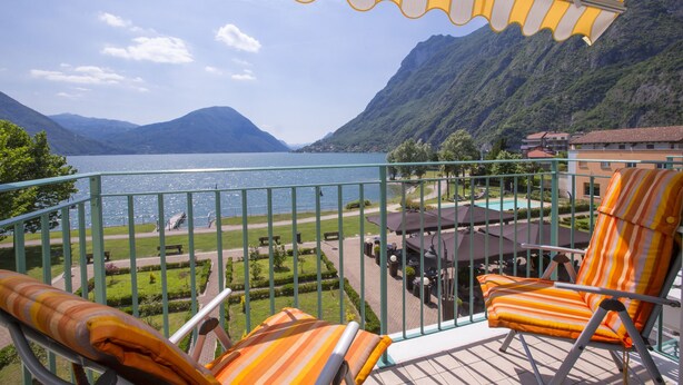 Een balkon met oranje en witte kussens in strepen, uitzicht op een meer, bergen en een zwembadgedeelte met parasols.