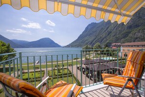 A balcony with striped orange and white cushions, a view of a lake, mountains, and a pool area with umbrellas. - Porto Letizia S12, Porlezza, Italy (Porlezza)