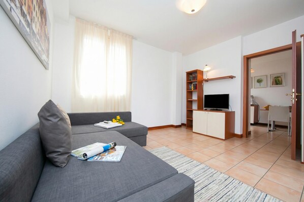 A modern living room with a grey sofa, a flat-screen TV, a bookshelf, and a dining area with a table and chairs. - Porto Letizia Begonia 6, Porlezza, Italy (Porlezza)