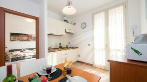A compact kitchen with white cabinets, a dining table set with plates, glasses, and a bottle. A clock and a fan are visible on the walls. - Porto Letizia Begonia 6, Porlezza, Italy (Porlezza)