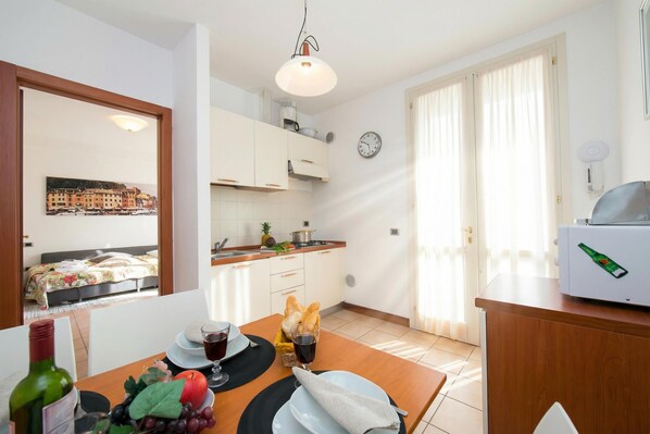 Cuina compacta amb armaris blancs, una taula de menjador preparada amb plats, gots i una ampolla. Hi ha un rellotge i un ventilador visibles a les parets.