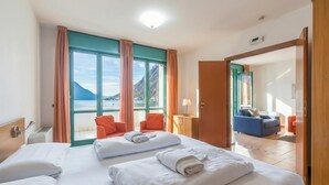 A hotel room with two beds, a view of the sea, and a balcony with orange chairs. - Porto Letizia P14, Porlezza, Italy (Porlezza)