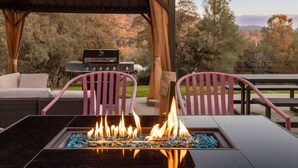 Un foyer avec un dessus en verre, entouré de chaises et agrémenté d’un barbecue, le tout installé dans un espace extérieur.