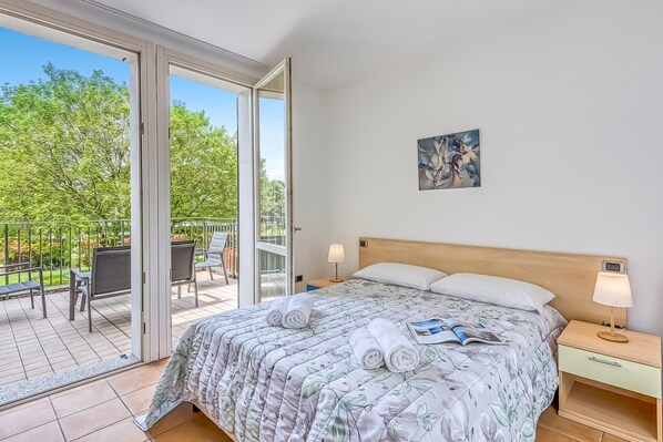A bedroom with a bed, a nightstand, a lamp, and a view of a balcony with chairs and a table.