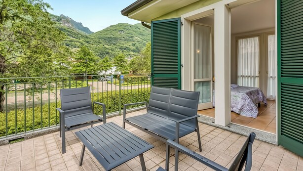 A balcony with two chairs and a table, a bed with a purple bedspread, and a mountain view. - Porto Letizia Rosa 2, Porlezza, Italy (Porlezza)