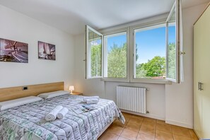 A bedroom with a bed, a radiator, a window with a view of trees, and two wall-mounted pictures.
