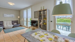 A living room with a sofa, TV, and a dining table with chairs. There is a bookshelf and a window with a view of the outside. - Porto Letizia Rosa 2, Porlezza, Italy (Porlezza)
