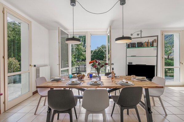 Uma sala de jantar com mesa de madeira posta para quatro pessoas, rodeada por cadeiras, com vista para o verde lá fora.
