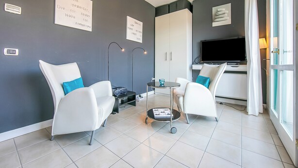 A modern living room with two white armchairs, a small round table, and a television. There are two wall-mounted lights, a cabinet, and a window with curtains. - Porto Letizia T1, Porlezza, Italy (Porlezza)