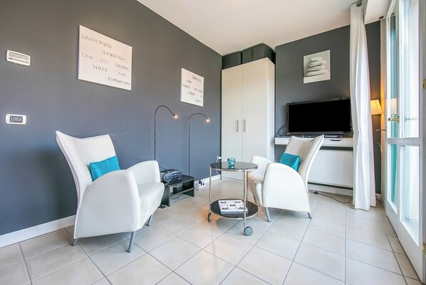 A modern living room with two white armchairs, a small round table, and a television. There are two wall-mounted lights, a cabinet, and a window with curtains.