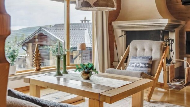 Ein gemütliches Wohnzimmer mit Holzbalkendecke, Kamin und großem Fenster mit Blick auf ein Haus.