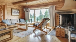 Ein gemütliches Blockhaus-Interieur mit Holzbalkendecke, Kamin, einer Couch, einem Schaukelstuhl und einem Esstisch.
