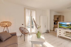 A modern living room with a sofa, a wicker chair, a coffee table with a vase, a television, and a kitchen area with cabinets and a microwave. - Porto Letizia Mimosa 3, Porlezza, Italy (Porlezza)