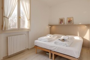 A bedroom with a double bed, a bedside table with a magazine, a window with curtains, a radiator, and two wall-mounted pictures.