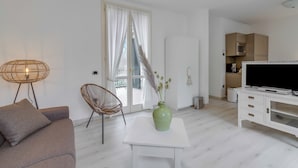 En moderne stue med en sofa, et sofabord med en vase, en lænestol, et fjernsyn og et køkkenområde med skabe og en mikrobølgeovn.