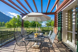 Een overdekt terras buiten met een eettafel, omringd door stoelen, en een parasol voor schaduw.