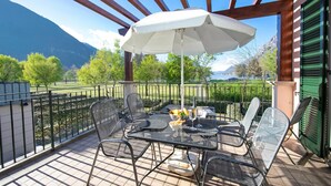 A terrace with a table and chairs, a white umbrella, and a view of mountains and a lake. - Porto Letizia Begonia 5, Porlezza, Italy (Porlezza)