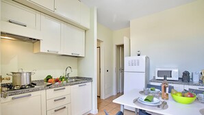 A modern kitchen with white cabinets, a gas stove, and a microwave. There is a dining table with chairs and a bowl of fruit. - Porto Letizia Begonia 5, Porlezza, Italy (Porlezza)