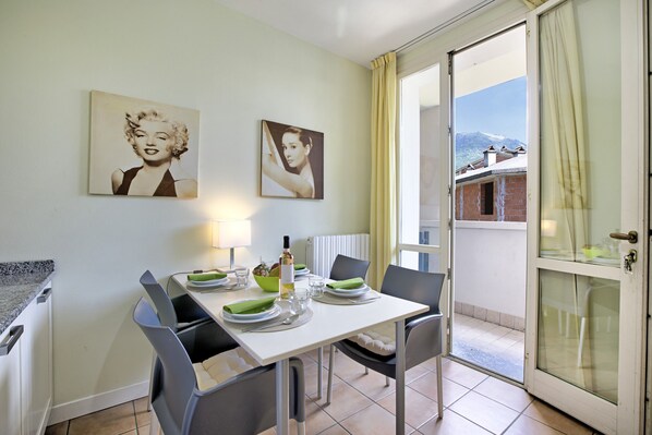 A dining area with a white table set for two, two framed pictures on the wall, and a view of a mountain through an open door. - Porto Letizia Begonia 5, Porlezza, Italy (Porlezza)