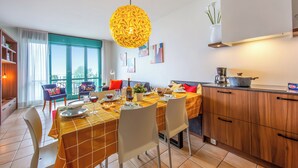 Un coin repas avec une table dressée pour quatre personnes, un espace cuisine équipé d’une cuisinière et d’un plan de travail, et une grande fenêtre avec des rideaux.