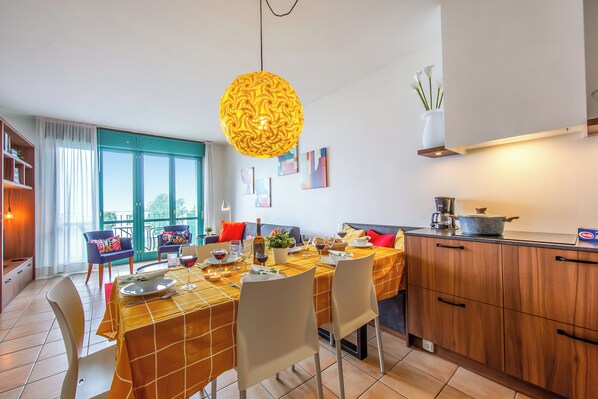 A dining area with a table set for four, a kitchen area with a stove and counter, and a large window with curtains. - Porto Letizia S7, Porlezza, Italy (Porlezza)