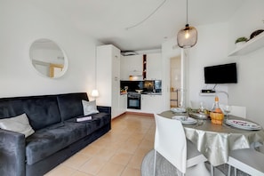 Een moderne woonkamer met een zwarte bank, een eettafel met witte stoelen, een keuken op de achtergrond en een wandgemonteerde televisie.
