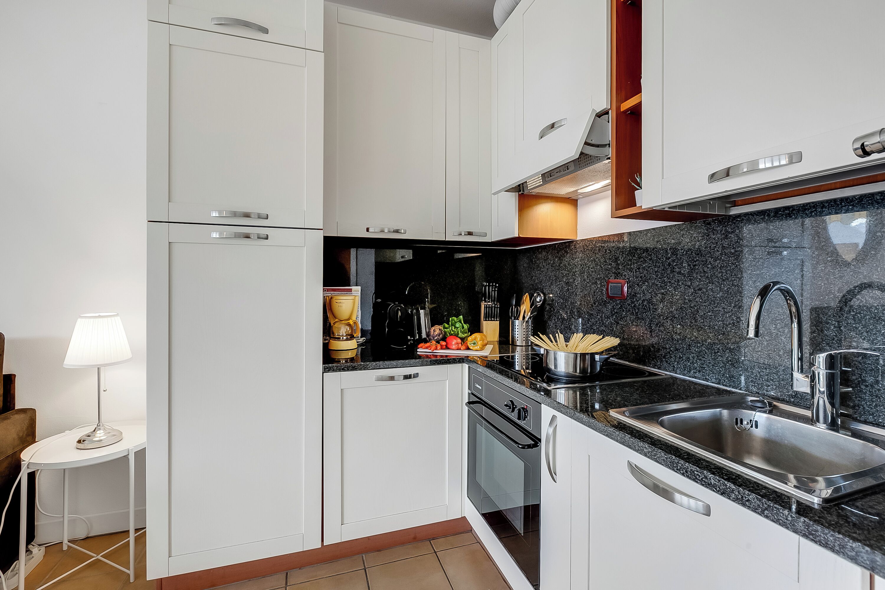 A modern kitchen with white cabinets, a granite countertop, stainless steel appliances, and a small dining area with a table and lamp.