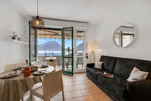 A dining area with a table set for two, a black sofa, and a view of the sea and mountains through open doors. - Porto Letizia P9, Porlezza, Italy (Porlezza)