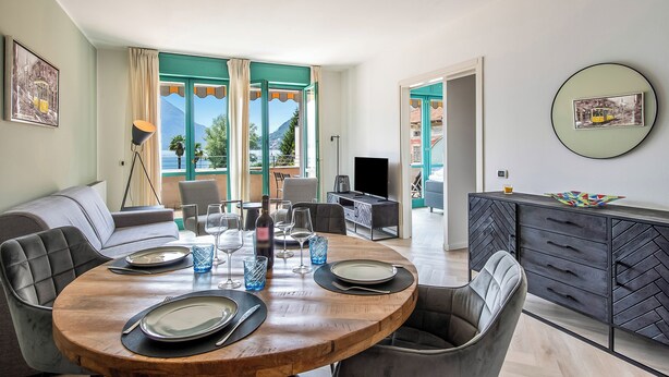 A dining area with a round wooden table set for four, a sofa, and a TV in the background. - Porto Letizia P7, Porlezza, Italy (Porlezza)