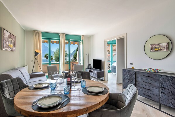 A dining area with a round wooden table set for four, a sofa, and a TV in the background. - Porto Letizia P7, Porlezza, Italy (Porlezza)