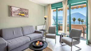 Uma sala de estar moderna com um sofá cinza, uma mesinha redonda com bebidas e uma cadeira. Um quadro emoldurado está pendurado na parede, e um abajur é visível.