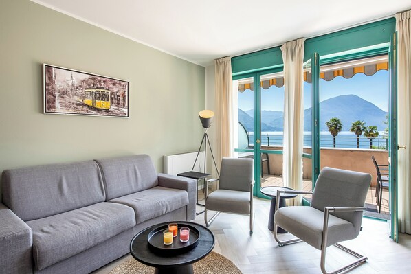 A modern living room with a grey sofa, a small round table with drinks, and a chair. A framed picture hangs on the wall, and a floor lamp is visible. - Porto Letizia P7, Porlezza, Italy (Porlezza)